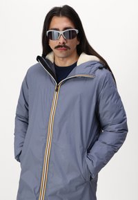 Giacca a vento blu con cappuccio foderato in morbido pile color crema e dettagli della zip multicolore; il modello indossa occhiali da sole grigi e una maglietta blu navy.