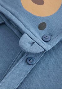 Blauter Stoff mit strukturiertem Finish, der ein großes braunes kreisförmiges Muster zeigt. Zwei dunkelblaue Knöpfe am Ausschnitt mit einem Riegel zum Sichern.