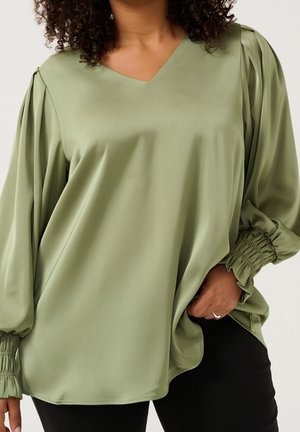 Femme portant un chemisier en satin vert sauge avec des poignets froncés et un col en V, main reposant dans la poche d'un pantalon noir.