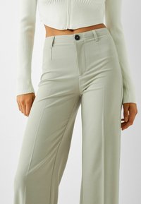 Pantalon vert clair avec une texture lisse, un design à jambes larges, des poches avant et un seul bouton à la taille.
