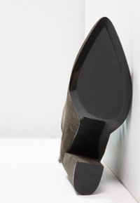 Botte en daim marron à bout pointu avec un talon bloc, présentant une semelle et un talon texturés noirs sur un fond blanc aux angles marqués.
