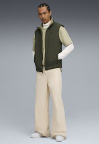Gilet senza maniche verde oliva, camicia leggera verde chiaro con texture, pantaloni larghi color crema e sneakers bianche. Presentato su uno sfondo sfumato.