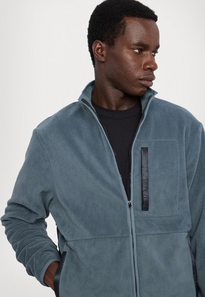 Veste en polaire de couleur bleu doux avec un col haut, une fermeture éclair sur le devant et une poche poitrine avec un accent en tissu noir. Texture douce, design décontracté.