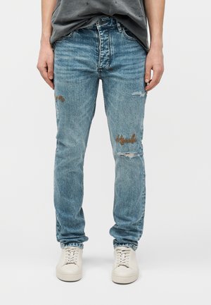 Personne portant un jean bleu clair usé avec des broderies sur les cuisses et des baskets blanches, debout devant un fond uni.