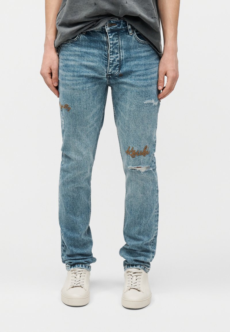 Άτομο που φοράει ανοιχτό μπλε distressed τζιν με κεντήματα στους μηρούς και λευκά αθλητικά παπούτσια, στέκεται μπροστά σε απλό φόντο.