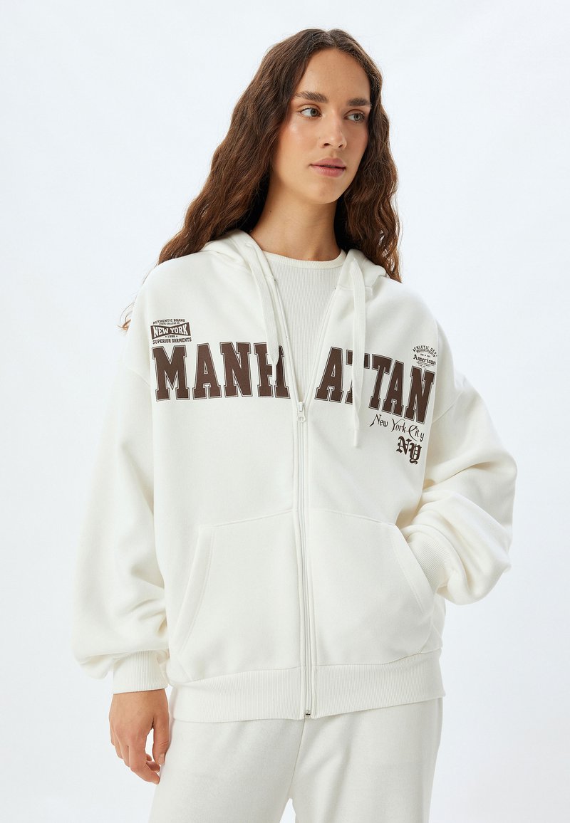 Weißer Reißverschluss-Hoodie aus weichem Material, auf dem die Aufschrift "MANHATTAN" in dunkelbraun auf der Vorderseite prangt und der über Kängurutaschen verfügt. Glatte Textur.