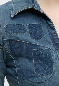 Veste en jean avec trois patchs visibles cousus sur la zone de la poitrine, de formes et nuances de bleu variées.