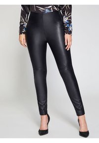 Femme portant des leggings en faux cuir noir et des escarpins noirs à talons aiguilles, les mains détendues le long du corps.