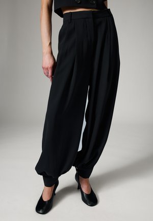 Un mannequin porte un pantalon noir taille haute à jambes larges, resserré par des fronces aux chevilles, et des talons noirs sur un fond de studio sobre.