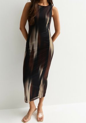 Vrouw die een mouwloze, getailleerde midijurk draagt met een abstract verticaal patroon in bruin, beige en zwart, gecombineerd met sandalen met doorzichtige bandjes en hak.