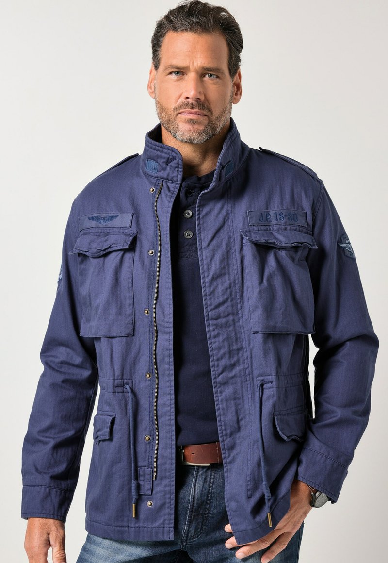 Laivastonsininen sotilastyylinen takki, valmistettu kankaasta, jossa on useita taskuja ja korkea kaulus. Kultasävyiset vetoketju ja metalliosat näkyvissä.