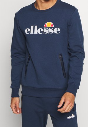 Mężczyzna ubrany w granatową bluzę ellesse i pasujące spodnie z naszywkami z logo oraz zameczkami na bocznych kieszeniach bluzy.