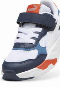 Baskets PUMA présentant un mélange de mesh blanc, de cuir bleu marine et d'accents orange. Le design comprend une sangle Velcro et une semelle texturée.