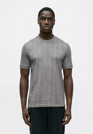 Mand står med fronten fremad, iført en grå kortærmet EA7 Emporio Armani T-shirt med lodrette logostriber, ensfarvet baggrund.