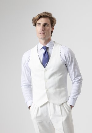 Joven con cabello castaño claro que lleva un chaleco y pantalones blancos, camisa de rayas y corbata azul con estampado, de pie con las manos en los bolsillos.