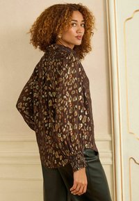 Blusa trasparente marrone con macchie leopardate metalliche, maniche lunghe a sbuffo e polsini arricciati. Indossata sopra pantaloni di pelle nera, evidenziando texture e design.