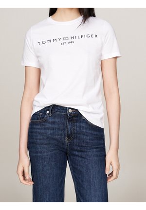 Kobieta ubrana w białą koszulkę Tommy Hilfiger i ciemnoniebieskie jeansy, stoi z rękami swobodnie opuszczonymi wzdłuż ciała.