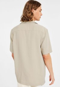 Beige Kurzarmhemd mit entspanntem Schnitt und einem einzelnen Rückensteg. Glatte Textur und dezenter Glanz, mit umgeschlagenem Kragen.