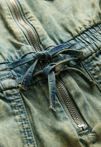 Denimová látka v svetlomodrej farbe s textúrovaným povrchom. Obsahuje strieborný zips, nastaviteľné viazanie v páse a zberaný detail.