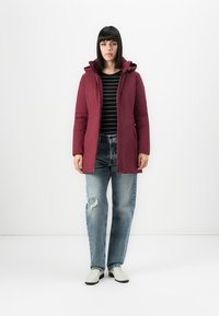 Vrouw gekleed in een bordeauxrode gewatteerde jas met capuchon, een zwart-en-witte gestreepte shirt, gescheurde blauwe spijkerbroek en witte schoenen, staande tegen een witte achtergrond.
