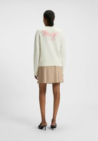 Jasnozielony sweter wykonany na drutach z różowym napisem "Hugo" na plecach, zestawiony z beżową spódnicą plisowaną i czarnymi klapkami na obcasie.