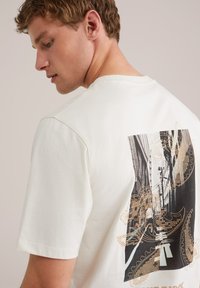 White cotton t-shirt featuring a black and white photograph print with gold paisley patterns on the back. Short sleeves and crew neck design.