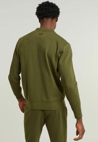 Felpa verde oliva con colletto a girocollo, maniche lunghe e polsini aderenti. Presenta un piccolo etichetta del marchio sul retro. Realizzata in tessuto morbido.