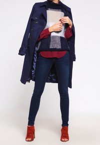 Person klädd i en marinblå kappa över en färgblockerad tröja, vinröd skjorta, mörka skinny jeans och orange öppna klackskor, stående mot en vit bakgrund.