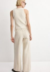 Mujer con cabello corto y rubio, lleva gafas de sol, top de punto crema sin mangas con encaje en el borde y pantalones anchos beige, de pie de espaldas.