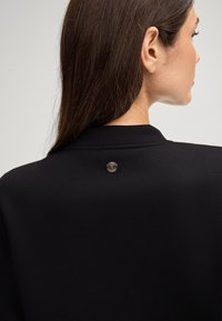 Vêtement noir avec une texture lisse, comportant un accent métallique rond à l'arrière près du col. Design simple et moderne.