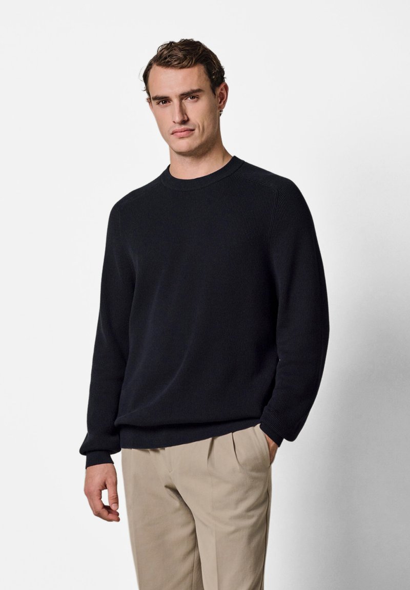 Mann mit kurzen braunen Haaren, der einen schwarzen Pullover mit langen Ärmeln und beige Hosen trägt und mit einer Hand in der Tasche vor einem weißen Hintergrund steht.