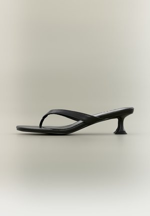 Sandale à talon haut en cuir noir avec une fine semelle et un talon sculpté en forme de petit chat, sur un fond beige uni.