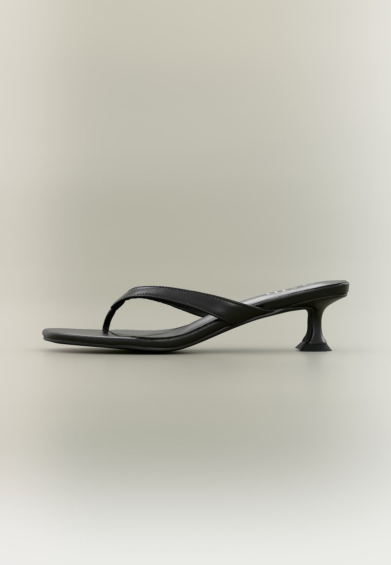 Sandale à talon haut en cuir noir avec une fine semelle et un talon sculpté en forme de petit chat, sur un fond beige uni.