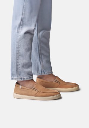 Jasnoniebieskie dżinsy w połączeniu z jasnobrązowymi butami wsuwanymi z białymi podeszwami i elastycznymi bocznymi panelami, pokazane na neutralnym tle.