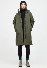 Olivgrüner wasserdichter Mantel mit Kapuze, durchgehendem Reißverschluss und Druckknöpfen. Kombiniert mit einem schwarzen Rollkragenpullover, Leggings und beigen Schuhen.