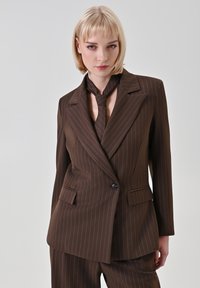 Abito marrone a righe con blazer doppiopetto, revers a incavo, tasche frontali e cravatta abbinata. Vestibilità su misura e tessuto liscio.