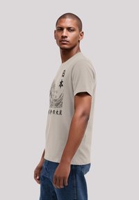 Lichtbeige t-shirt met een zwart golfgrafiek en Japanse tekst. Korte mouwen, ronde hals, gemaakt van zachte stof, comfortabele pasvorm. Spijkerbroek.