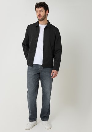Homme portant une veste noire, une chemise blanche, un jean bleu et des baskets blanches, debout devant un fond clair uni.