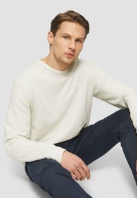 Lichtcrème sweatshirt met een ronde hals en geribde manchetten, gedragen over donkerblauwe broeken. Minimalistisch ontwerp met een subtiele merkdetails.