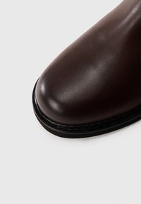 Bota corta de cuero marrón con punta redonda, textura lisa y suela de goma negra. Se aprecia costura contrastante a lo largo del borde.