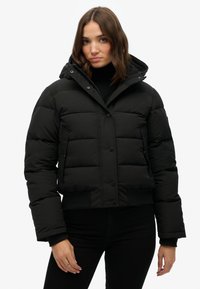 Chaqueta acolchada negra con diseño corto, cierre de botones, bolsillos laterales y capucha desmontable, con un acabado suave y texturizado.