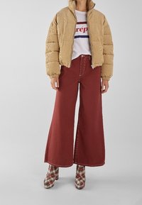 Chaqueta acolchada beige con textura acanalada, camiseta gráfica blanca, pantalones anchos burdeos y botines a cuadros con tacón.
