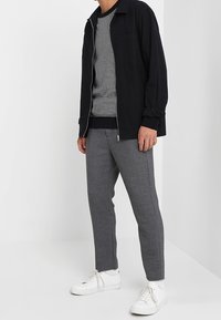 Giacca nera con zip sopra un maglione a righe grigie e bianche, abbinata a pantaloni grigi e sneakers bianche. Caratteristiche includono maniche a coste e tasche.