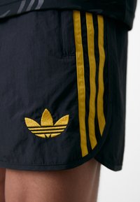Shorts de sport noirs avec trois rayures verticales jaunes sur le côté et un logo Adidas en jaune brodé près de l'ourlet.