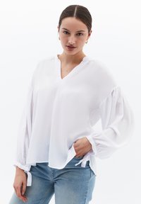 Blusa bianca con scollo a V, maniche lunghe morbide e ampie, con polsini arricciati. Tessuto morbido con una silhouette rilassata. Abbinata a jeans azzurri chiari.