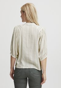 Blusa de un tono beige claro con rayas verticales, mangas 3/4 abullonadas y un corte holgado. Textura suave y detalle de patrón sutil en la tela.