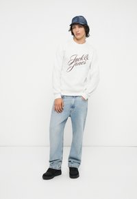 Fehér pulóver "Jack & Jones" bordó írással, világoskék, laza fazonú farmerrel és fekete sportcipővel párosítva. Akryl sapka.