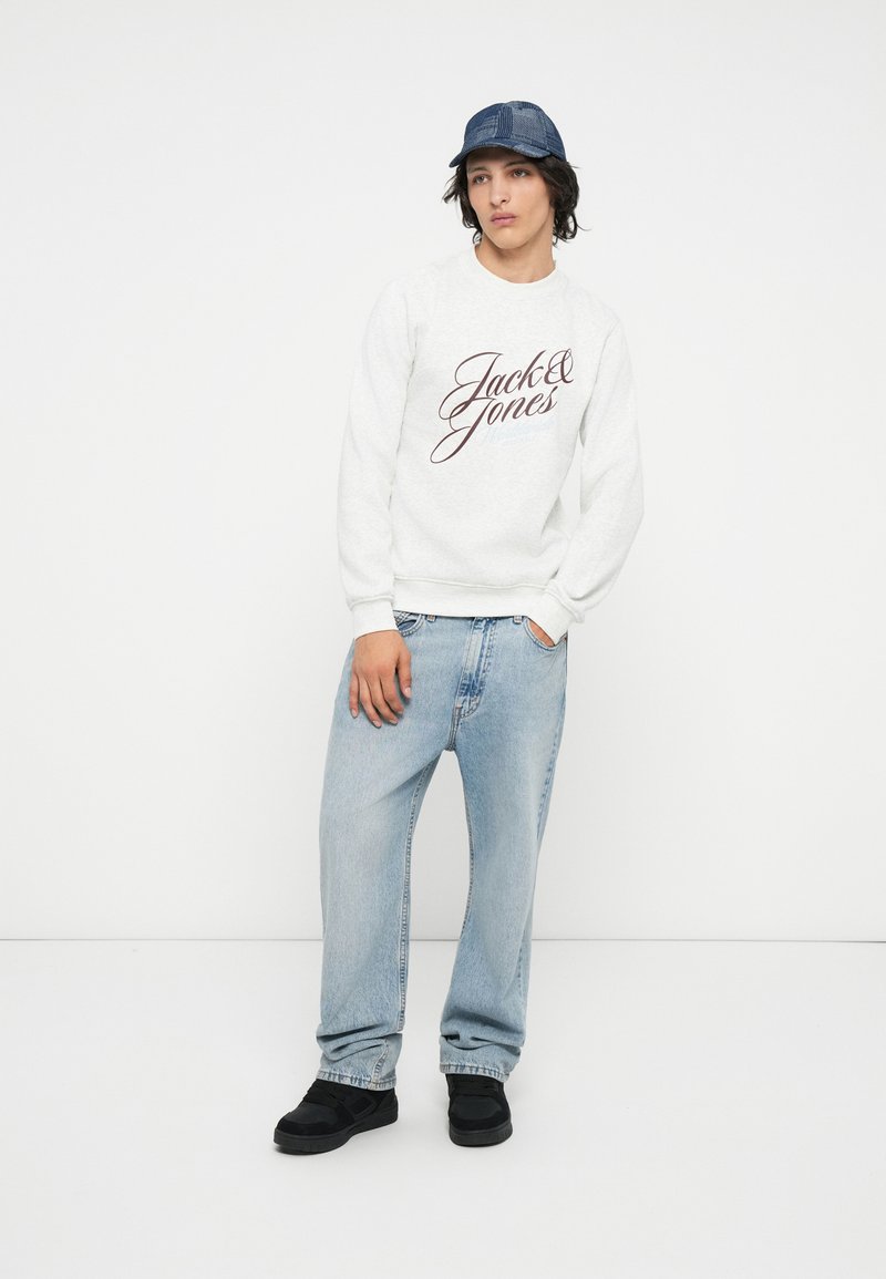 Sudadera blanca con "Jack & Jones" en letras burdeos, combinada con unos vaqueros de corte relajado en azul claro y zapatillas negras. Gorra de acrílico.