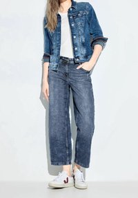 Chaqueta de mezclilla azul con botones frontales y bolsillos, combinada con unos vaqueros oscuros de corte ancho y zapatillas blancas con detalles rojos.