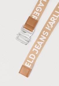 Cinto de tecido bege com texto trançado em branco, apresentando uma fivela de metal e acabamentos em couro nas extremidades. Design robusto, textura têxtil.